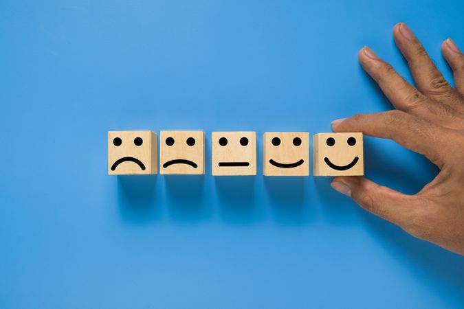male hand put wooden blocks with five different emoji expression from sad to happy isolated on blue background. rating scale satisfaction feedback concept