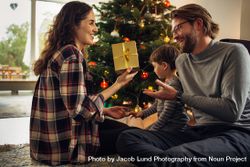 Couple sitting by Christmas tree with their son exchanging gifts at home 47L9Ob