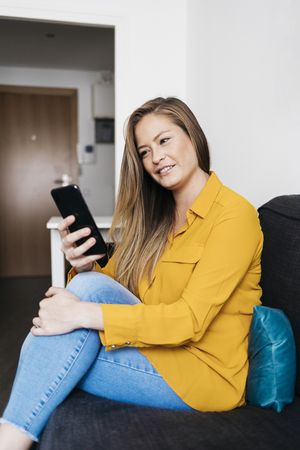 Beautiful adult female sitting on sofa while using smartphone