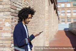 Side view of Black man looking down at his smartphone while leaning on a brick wall outdoors on sunny day 5lVRAe