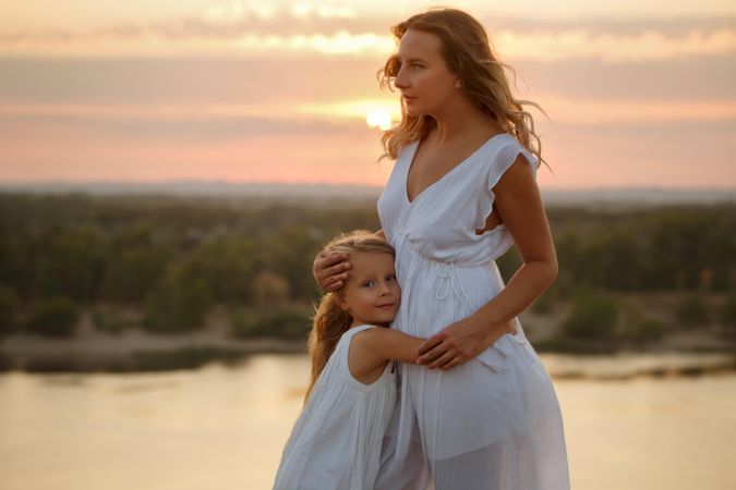 Woman and child in dresses hugging in front of lake during beautiful sunset