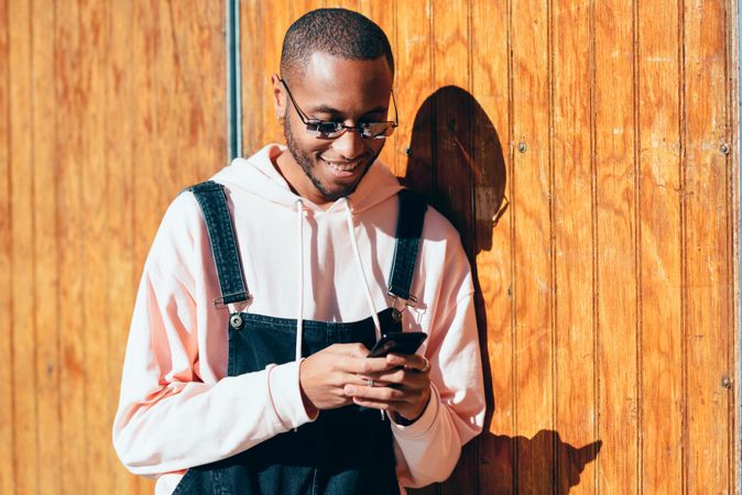 Male wearing overalls and smiling and checking phone standing by a wooden wall