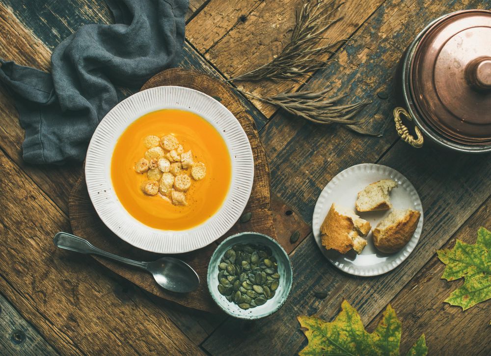 Pumpkin soup with croutons, bread, seeds, pot, spoon on wooden table with leaves