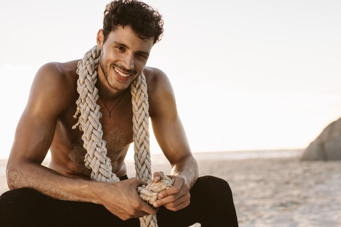Shirtless man relaxing after workout at a beach with a battle rope on his neck