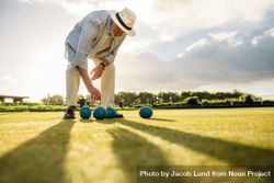Mature man in hat bending down to pick a boules standing in a playground 5lmNm4