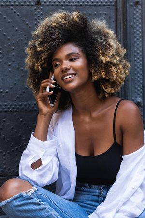 Delighted Black woman speaking on smartphone while sitting near building