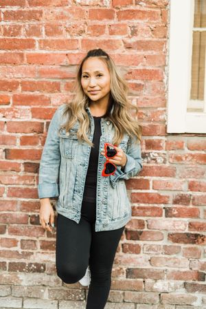Woman in denim jacket holding red framed sunglasses leaning against red brick wall