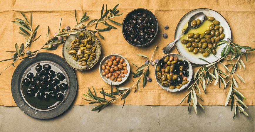 Olives in bowls with branches, on concrete with beige table linen, wide composition