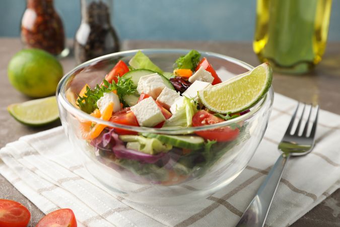 Glass bowl with salad, towel and spices on grey background, space for text