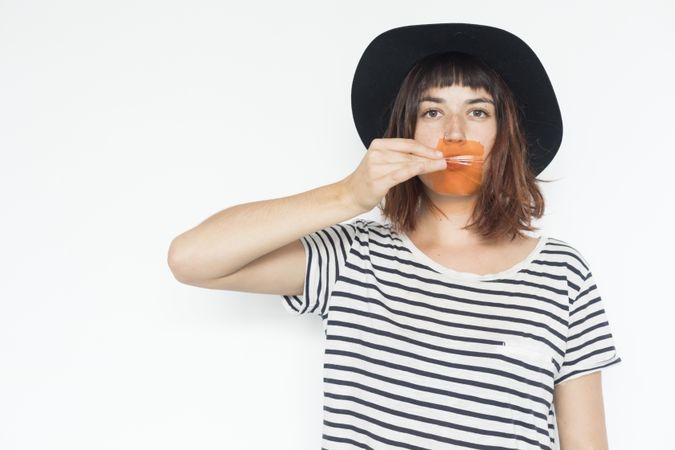 Female in striped shirt and felt hat holding transparent orange material to mouth