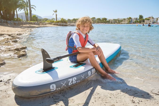 Cheerful boy sitting on paddleboard on sandy beach