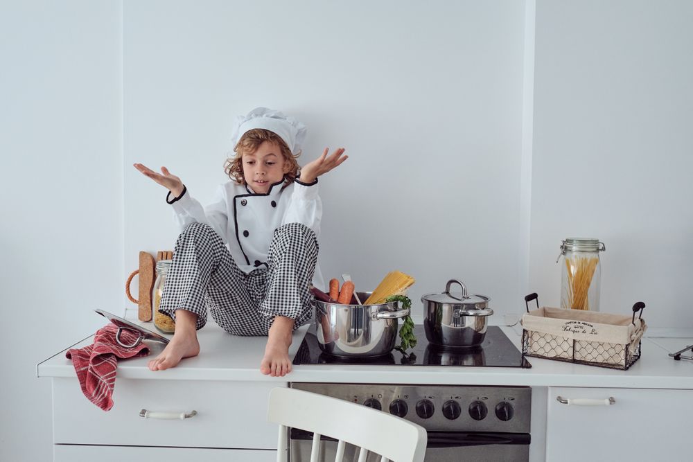 Bewildered preteen boy in chef costume sitting on kitchen counte