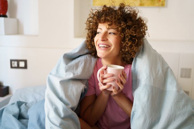 Smiling woman relaxing under blue sheet in bed with cup of coffee