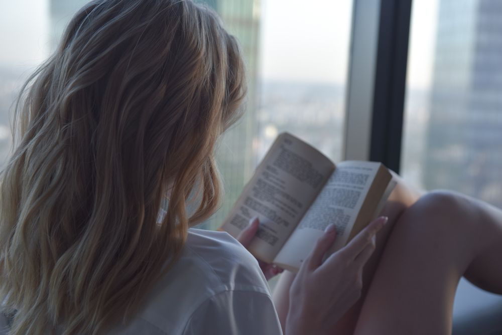 Back view of woman reading book sitting beside window
