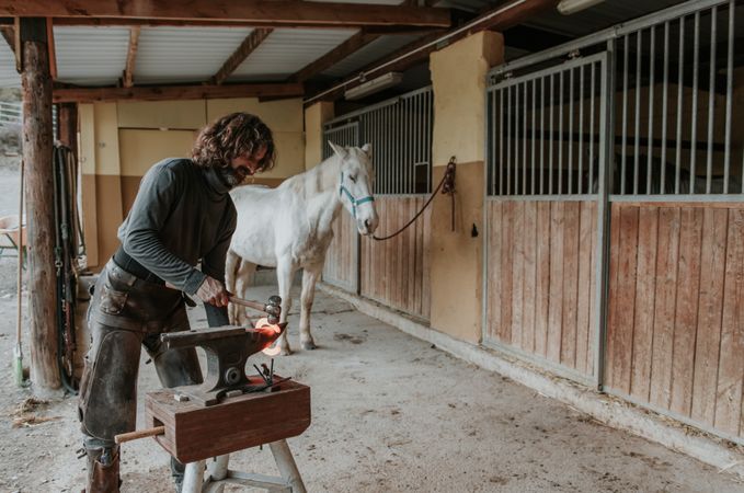 Blacksmith forging horseshoe near stable
