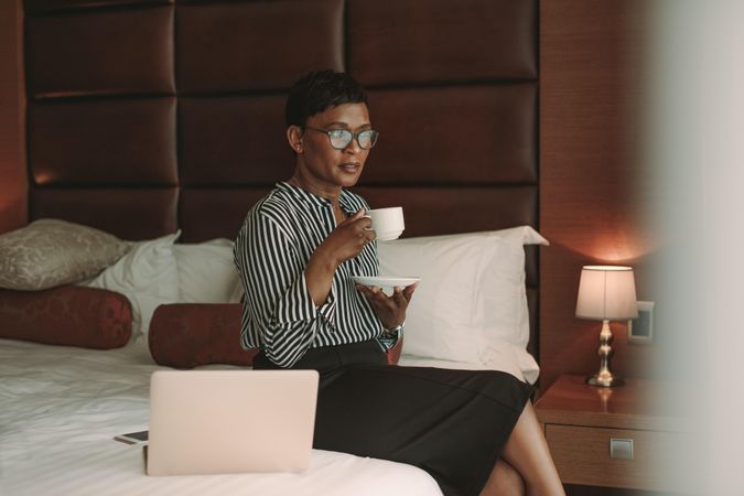 Woman in formal clothes sitting on bed in hotel room and having a cup of coffee