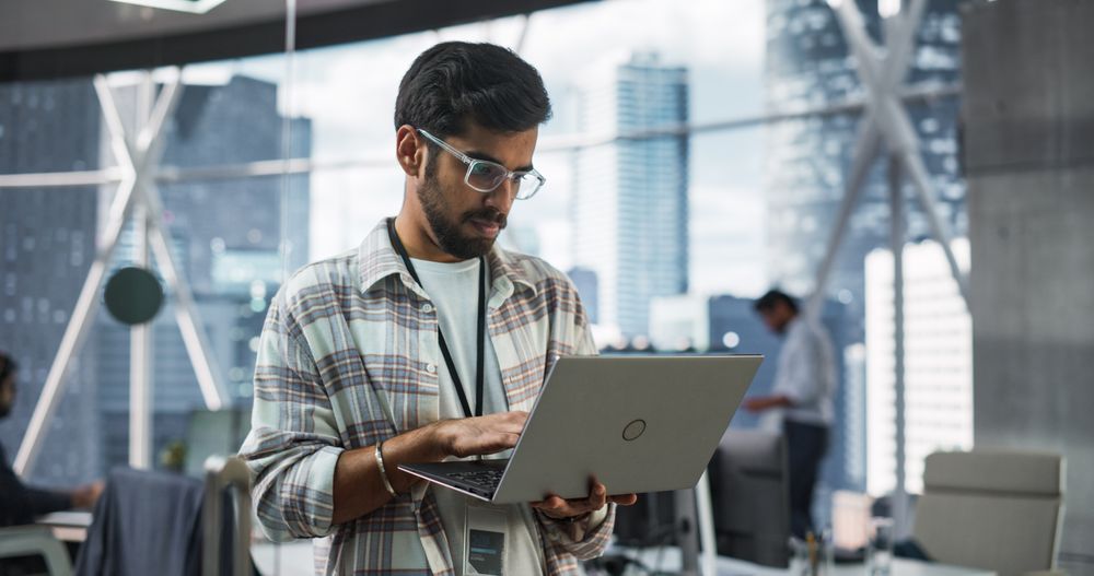 Indian Male Business Analyst Standing with Laptop Computer in a Modern Office.