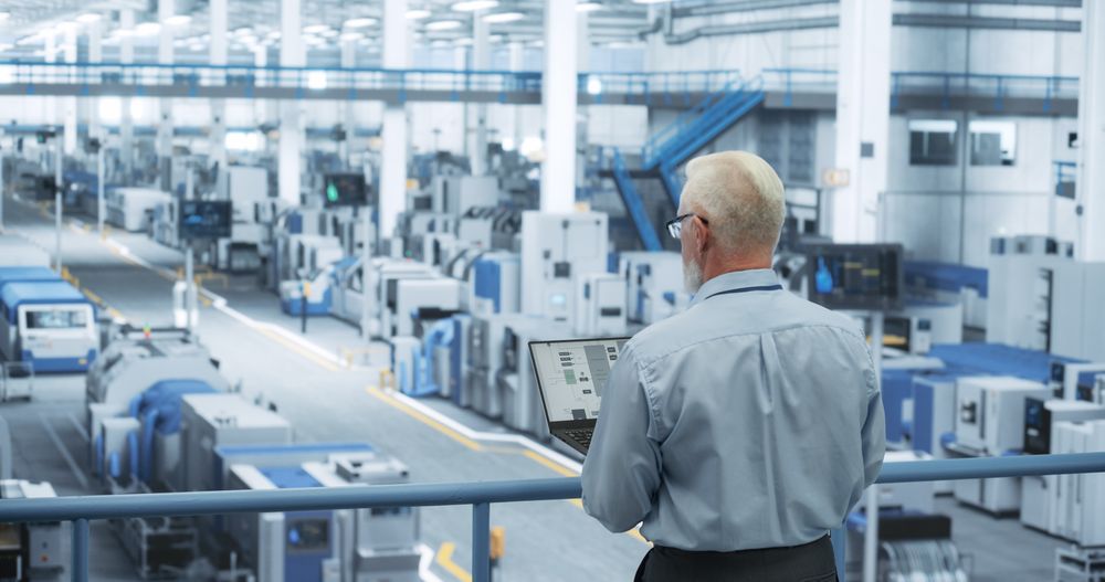 Portrait of an Adult Engineer Standing on a Platform, Using Laptop Computer at an Electronics Factory. Technician Making Technical Upgrades Online Over an Internet Software