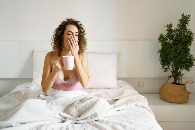 Woman yawning as she sips her coffee in bed