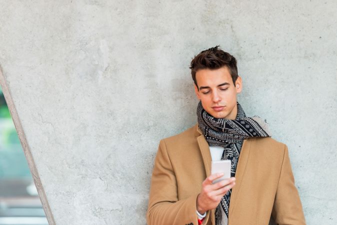 Young man wearing winter coat looking at phone screen against cement pillar