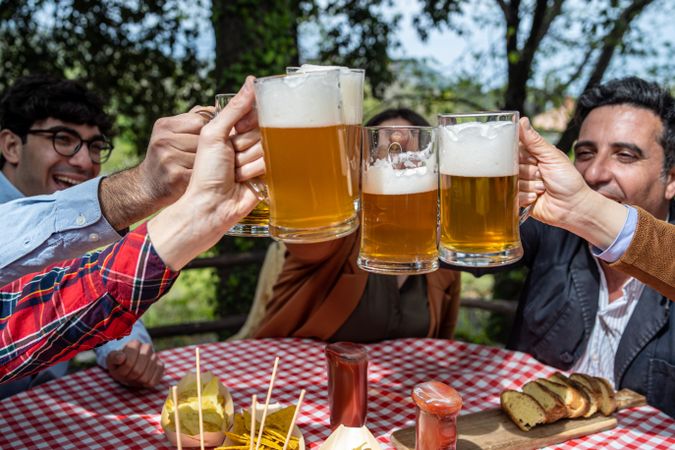 Group of happy friends celebrate friendship toasting with beer mugs sitting outdoors at picnic table