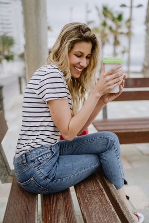 Happy woman with disposable cup of coffee sitting on street bench