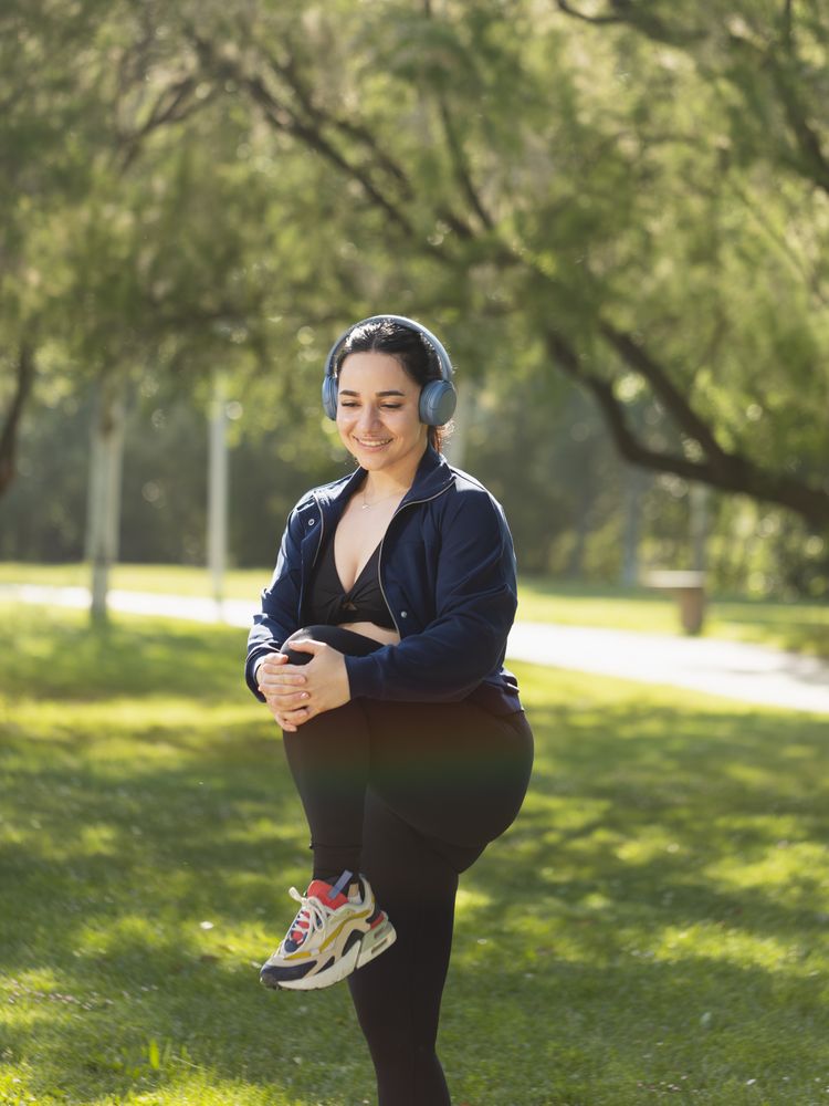 A young woman performs exercises and stretches of legs, biceps and buttocks in the morning in the park, while listening to music