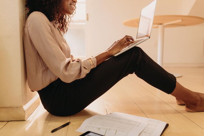 Businesswoman sitting on floor at home working on laptop with business papers by her side