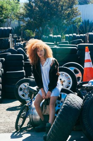 Full length portrait of young woman in light dress and brown fur coat sitting on tires