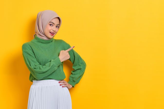 Smiling woman in headscarf pointing up yellow copy space