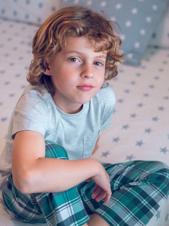 Adorable boy with curly hair and nightwear sitting on bed