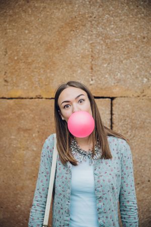 Woman standing outside in front of stone wall blowing bubble with pink gum