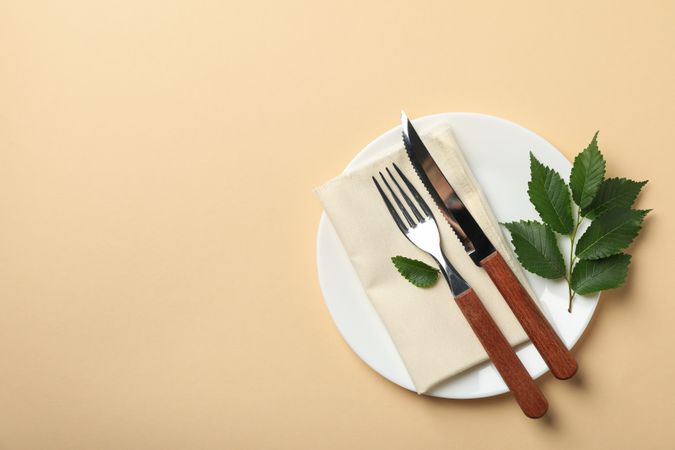 Plate with napkin, cutlery and leaf on beige background, top view