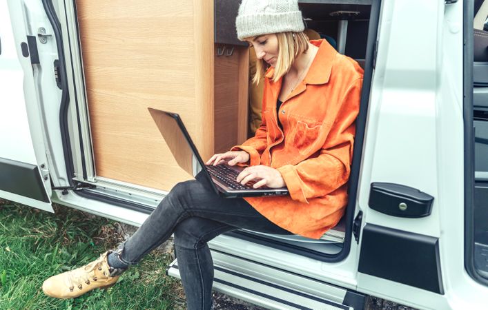 Female in jacket sitting on step of van with laptop