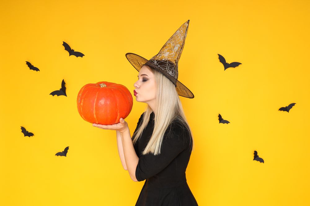 A woman in a wizard's hat kisses a pumpkin, on a yellow background