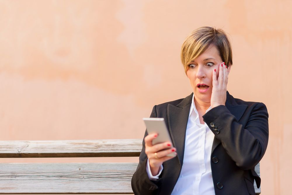 Shocked female in blazer sitting on bench checking phone in front of peach wall