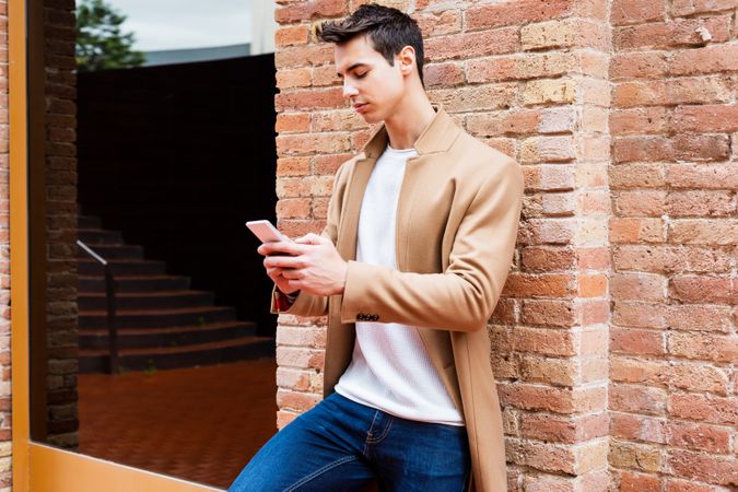 Male in camel coat texting on phone while leaning on brick wall