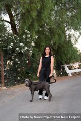 Full length portrait of a woman in casual attire standing in the street with her dog on a leash 48BVK0
