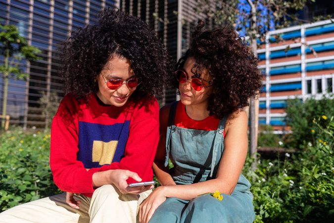 Two female friends sitting on bench outside checking phone together