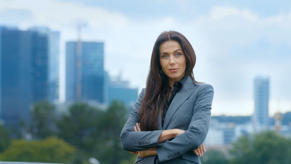 Successful Business Woman Crosses Arms and Smiles in the Background Skyscrapers and Business Centre of the Big City is Visible.
