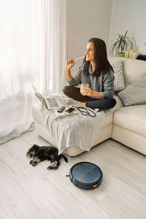 Smiling female sitting on sofa near sleeping dog and robotic vac