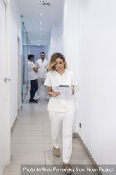 Female doctor walking in clinical corridor while holding a medical folder with medical team in the background 4MQ1z4
