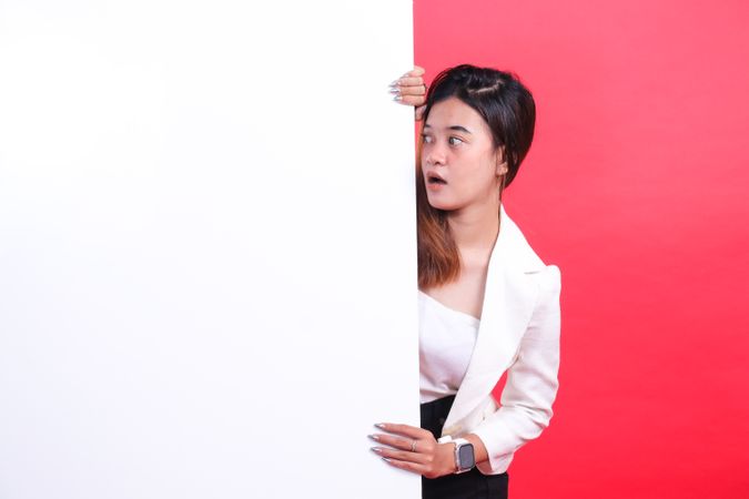 A woman wearing a jacket and a skirt is holding a large cardboard while looking to the side, isolated on a red background