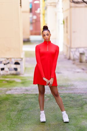 Serious woman in casual red dress standing in alley on sunny day