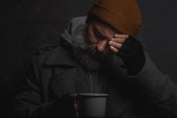Sad homeless man with a gray beard holding a cup of hot tea to warm himself on a cold night