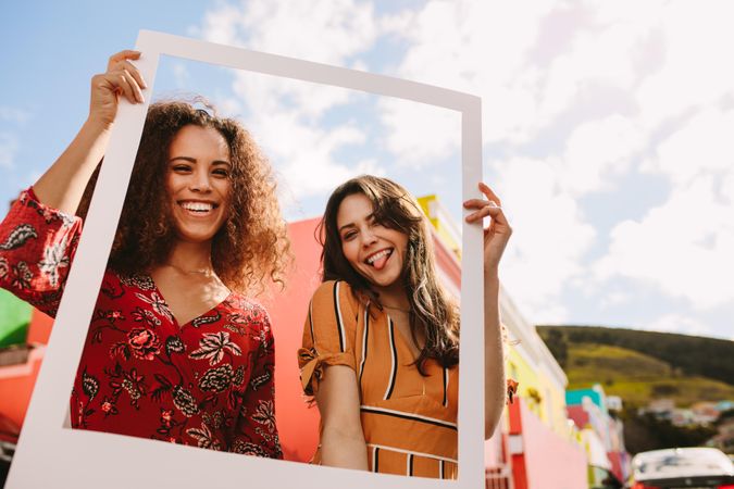 Two smiling women standing together and holding empty frame outdoors
