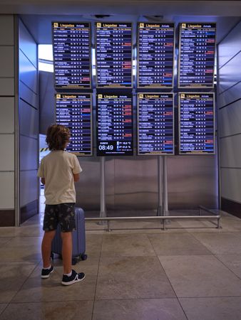 Unrecognizable boy with blue suitcase looking at arrival boards