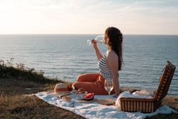 Woman with glass of drink on picnic mat looking at sea and mountains