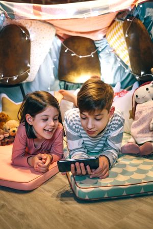 Happy little girl laughing while watch video on streaming platform in cell with boy lying over mat