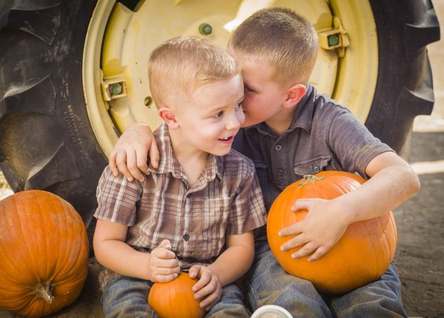 Two Boys Sitting Against Tractor Tire Holding Pumpkins Whispering Secrets